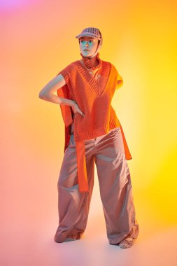 Tasarımcı örgüsü. Geniş paçalı pançoyla poz veren şık bir manken, atölyede örgü ören panço ve karışık renkli ışıkta örgü ören bir şapka. Parlak renkler. Yeni moda yarı sezon koleksiyonu. 