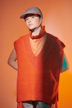 Yarı sezonluk özel tasarım koleksiyonu. Örgü giyim tarzı. Geniş paçalı pançoyla poz veren şık bir manken, atölyede örgü ören panço ve karışık renkli ışıkta örgü ören bir şapka. Parlak renkler.