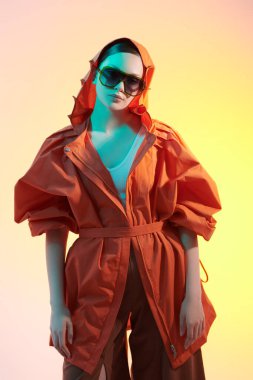 Yaz koleksiyonu. Geniş kollu ve geniş bacaklı kahverengi pantolonlu şık turuncu ceketli bir manken kız. Stüdyo görüntüsü sarı arkaplan ve karışık renkli ışıklandırma. 