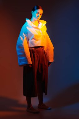 Moda ve moda tarzında. Güzel manken kız geniş şekilli şık kıyafetler içinde poz veriyor. Stüdyo karman çorman bir ışıkta koyu bir arkaplan karşısında. Parlak renkler.