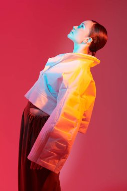 Moda ve moda tarzında. Güzel manken kız geniş şekilli şık kıyafetler içinde poz veriyor. Kırmızı arkaplanda karışık renk ışıklarıyla çekilmiş bir stüdyo çekimi. Parlak renkler.