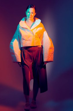 Moda ve moda tarzında. Güzel manken kız geniş şekilli şık kıyafetler içinde poz veriyor. Stüdyo karman çorman bir ışıkta koyu bir arkaplan karşısında. Parlak renkler.