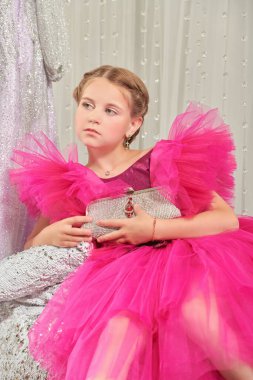 Lüks bir fuşya elbisesi içinde poz veren ve gece elbiselerinden oluşan bir salonda gümüş bir el çantasıyla küçük tatlı bir bayanın portresi. Akşam saç stili. Çocuklar için Yüksek Moda.