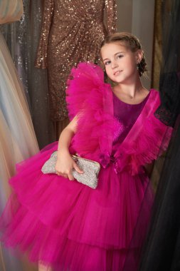 Lüks bir fuşya elbisesi içinde poz veren ve gece elbiselerinden oluşan bir salonda gümüş bir el çantasıyla küçük tatlı bir bayanın portresi. Akşam saç stili. Çocuklar için Yüksek Moda.