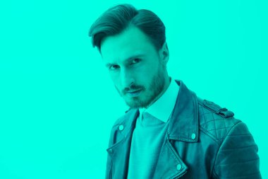 Siyah deri ceketli ve klasik elbiseli ciddi bir adam parlak turkuaz ışıkta duruyor ve kameraya ciddi ciddi bakıyor. Duygular, duygular, psikoloji. Metin için yer.