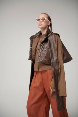 Özel tasarım koleksiyonu. Tasarımcı kahverengi deri ceket, bluz ve turuncu geniş bacak pantolonuyla poz veren güzel bir manken kız. Stüdyo görüntüsü gri arka planda. Şık güneş gözlükleri..