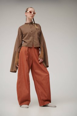 Eğilimler. Özel tasarım koleksiyonu. Tasarımcı kahverengi gömleği, örgü eldivenleri ve turuncu geniş paça pantolonuyla poz veren güzel bir manken kız. Stüdyo görüntüsü gri arka planda. Şık güneş gözlükleri..