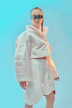 Yüksek moda ve fütüristik tarz. Beyaz desenli ceket, beyaz şort ve mavi stüdyo arka planında fütüristik aynalı güneş gözlüğü olan güzel bir kadın model poz veriyor. Gözlük stili.