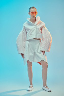 Yüksek moda. Beyaz ceketli, beyaz pantolonlu ve mavi stüdyo arka planında büyük turuncu gözlüklü güzel bir kadın model poz veriyor. Gelecekçi tarzı. Gözlük stili.