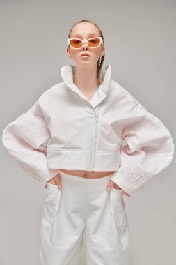 Yüksek moda. Gri stüdyo arka planında beyaz tasarımcı kıyafetleri ve zarif güneş gözlükleriyle güzel bir kadın model poz veriyor. Saç stili örgülü. Gözlük Moda.