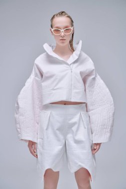 Yüksek moda. Gri stüdyo arka planında beyaz tasarımcı kıyafetleri ve zarif güneş gözlükleriyle güzel bir kadın model poz veriyor. Saç stili örgülü. Gözlük Moda.