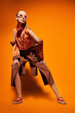 Moda fotoğrafı. Turuncu stüdyo arka planında yaz koleksiyonundan tasarımcı kıyafetleriyle poz veren güzel bir manken kız. Tam boy portre.