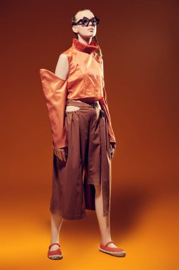 Yaz için özel tasarım koleksiyonu. Moda mankeni kız şık elmas şekilli gözlüklerle poz veriyor turuncu kolları sökülebilir ve kahverengi geniş bacaklı bir bluzu turuncu stüdyo arka planında.. 
