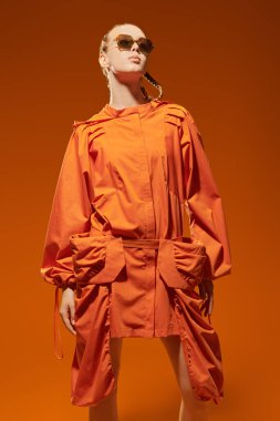 Yüksek moda çekimi. Portakal rengi pelerinli ve son moda güneş gözlüklü şık bir manken kız turuncu stüdyo arka planında poz veriyor. Yaz koleksiyonu. Örgülü saç stili.