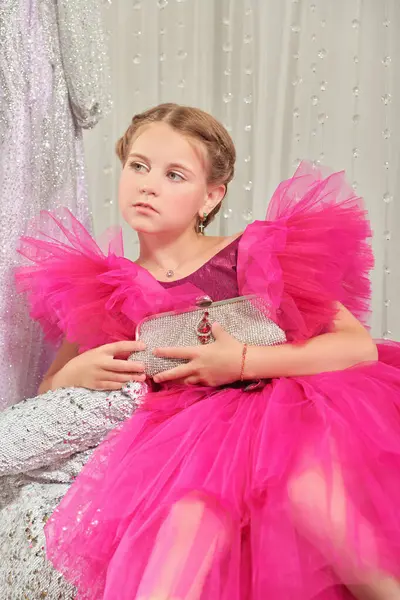 Lüks bir fuşya elbisesi içinde poz veren ve gece elbiselerinden oluşan bir salonda gümüş bir el çantasıyla küçük tatlı bir bayanın portresi. Akşam saç stili. Çocuklar için Yüksek Moda.