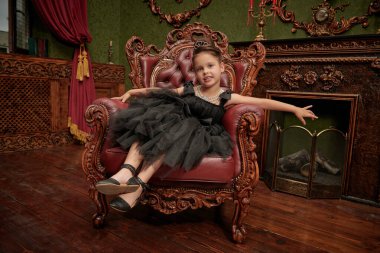 Kabarık siyah bir elbise giymiş, akşam saç stili ve takıları klasik bir koltuk ve gülümsemeyle süslenmiş güzel bir kızın tam boy portresi. Lüks daireler. Kızlar için akşam modası.