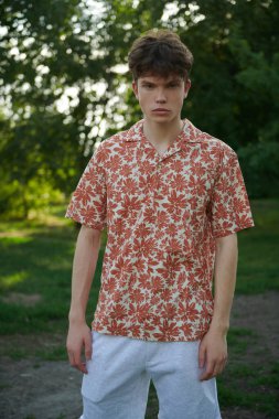 Yazlık gömlekli ve şortlu yakışıklı bir gencin portresi bir yaz parkındaki ağaçların arka planına karşı. İnsanlar, hayat anları, duygular. Yaz zamanı..