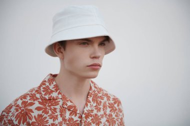 İnsanlar, hayat anları, duygular. Beyaz bir duvara karşı beyaz panama ve yazlık gömlekli modern bir gencin portresi. Sokak fotoğrafı. Yaz modası.