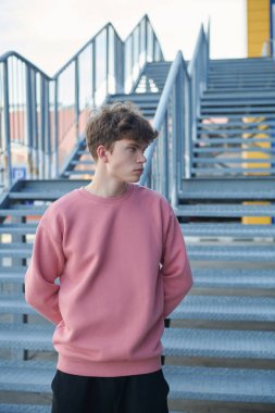 Modern bir genç manken, alışveriş merkezinin yanındaki metal bir merdivende pembe bir sweatshirt ve siyah şortla poz veriyor. Şehir tarzı. Gençlik modası.