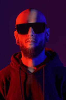 Dudağında piercing olan, siyah güneş gözlüğü ve siyah kapüşonlu kapüşonlu vahşi, sakallı bir adam. Stüdyo görüntüsü koyu mor arkaplanda karışık kırmızı ve mavi ışıkta.