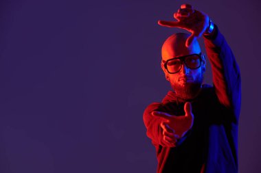 Kel, sakallı, dudak piercingli, siyah gömlekli ve büyük gözlüklü vahşi bir adamın karışık renkli portresi. Metin için yeri olan stüdyonun mor arkaplanı.