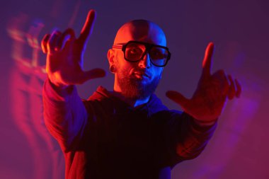 Kel, sakallı, dudak piercingli, siyah tişörtlü ve büyük gözlüklü vahşi bir adamın karışık renkli görüntüsü. Stüdyo mor arkaplan.