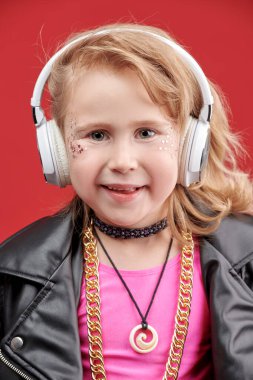 Parlak makyajlı, siyah deri ceketli ve kulaklıklı, kırmızı stüdyo arka planındaki pop ve rock müzisyeni gibi şirin bir kızın portresi. Çocuk modası. Küçük DJ kız.