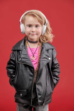 Parlak makyajlı, siyah deri ceketli ve kulaklıklı, kırmızı stüdyo arka planındaki pop ve rock müzisyeni gibi şirin bir kızın portresi. Çocuk modası. Küçük DJ kız.