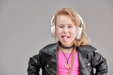 Parlak makyajlı, modaya uygun kıyafetleri ve aksesuarları olan sevimli bir kız ve rock müzisyeni kulaklıkla müzik dinliyor. Gri stüdyo geçmişi. Çocuk modası. DJ çocuk.