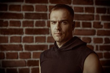 Siyah kapüşonlu güçlü bir adamın portresi, kameraya özgüvenle bakıyor. Arka planda tuğla duvar var. Halk konsepti. Erkekler, erkeklik ve güç.. 