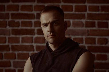 Erkekler, erkeklik ve güç. Siyah kapüşonlu güçlü bir adamın portresi, odaklanmış ve yoğun bir şekilde kameraya bakıyor. Arka planda tuğla duvar var. Halk konsepti.