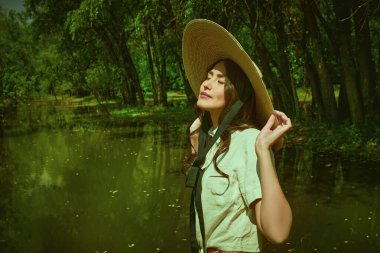 Geniş kenarlı hasır şapkalı, romantik bir Asyalı kız yaz parkındaki güzel bir göletin yanında hafif bir gülümsemeyle duruyor.. 