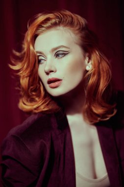 Pahalı kadife ceketli, loş ışıklı bir odada kızıl saçlı çekici bir genç kadın. Moda ve kadınlık. Kadın güzelliği ve tarzı. Oklarla ve saç stiliyle zarif bir makyaj..