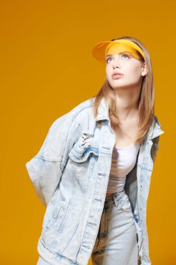 Denim gençlik tarzı. Kot ceketli güzel sarışın bir kızın portresi ve sarı bir stüdyo arka planında şık sarı bir güneşlik. Yaz modası. Spor kıyafetleri..