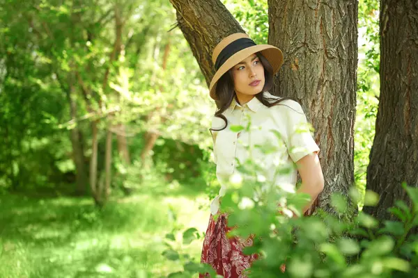 Yaz mutluluğu ve kaygısız ruh hali. Hasır şapkalı güzel bir Asyalı kız güneşli bir günde parkta yürüyor ve sıcak güneş ışınlarının tadını çıkarıyor..