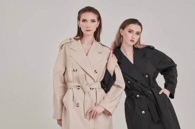 Beyaz bir stüdyo arka planında moda trençkotlarla poz veren iki şık güzel kız mankeni. Kadın modası ve güzelliği.