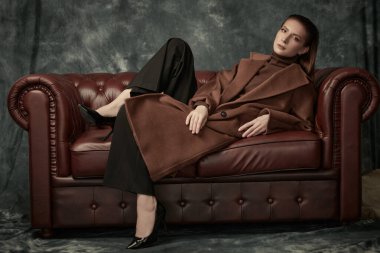 Çekici, kendine güvenen bir kadın deri koltukta poz veriyor moda keçeli ceketi, siyah pantolonu ve yüksek topuklu ayakkabıları var. Kadın modası.