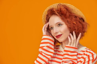 Kıvırcık kızıl saçlı güzel bir kız, hasır şapka giymiş ve sarı stüdyo arka planında turuncu çizgileri olan uzun kollu bir kız. İnsanların duyguları. Yaz modası ve güzelliği.