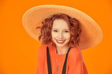 Portakal rengi stüdyo arka planında geniş kenarlı hasır şapkalı parlak kırmızı kıvırcık saçlı sevimli romantik bir kızın portresi. Mutlu insanlar, olumlu duygular. Yaz modası ve güzelliği. 