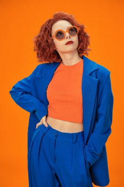 Parlak renkler. Çekici kıvırcık saçlı, parlak mavi, turuncu giysiler ve güneş gözlükleriyle gülümseyen kendine güvenen güzel bir kızın portresi. Stüdyo turuncu arka plan.