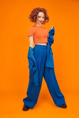 Parlak kırmızı kıvırcık saçlı, mavi ve turuncu elbiseli çekici, neşeli manken kızın tam boy fotoğrafı. Parlak renkler. Stüdyo turuncu arka plan. Yaz modası. 