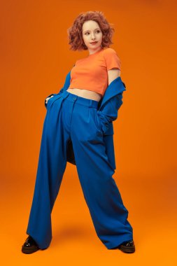 Parlak kırmızı kıvırcık saçlı, mavi ve turuncu elbiseli çekici, neşeli model kızın tam boy portresi. Parlak renkler. Stüdyo turuncu arka plan. Yaz modası. 