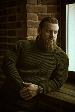 Tuğla bir duvarın arkasında sakince ve düşünceli bir şekilde duran örgü örülü süveter giyen yakışıklı bir sakallı adamın portresi..