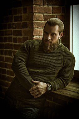 Tuğla bir duvarın arkasında sakince ve düşünceli bir şekilde duran örgü örülü süveter giyen yakışıklı bir sakallı adamın portresi..