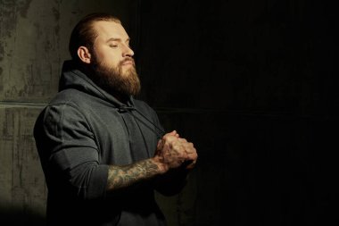 Erkeklik ve fiziksel güç. Erkek güzeli. Siyah kapüşonlu, iri yarı, sakallı bir adamın portresi. Arka planda beton bir duvar var. Boşluğu kopyala. 