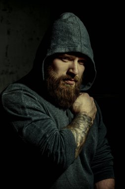 Erkeklik ve fiziksel güç kavramı. Erkek güzeli. Siyah kapüşonlu, iri yarı, sakallı bir adamın portresi. Arka planda beton bir duvar var.. 