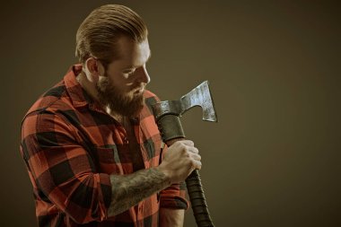 Erkek güzelliği ve erkeklik. Elinde baltası olan, ekose gömlekli, vahşi sakallı bir adamın portresi. Fotokopi alanı olan koyu kahverengi bir stüdyo. Berber dükkanı reklamı.