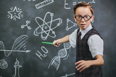 Okul üniformalı, gözlüklü ve kameraya şaşkınlıkla bakan zeki bir çocuğun portresi. Bilimsel çizimleri olan siyah bir karatahta. Eğitim, öğretmenlik. Genç bilim adamı.