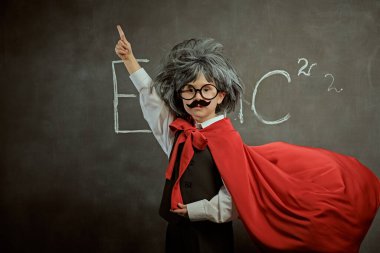 Eğitim ve erken gelişim. Küçük Bilim adamları. Gözlüklü, bıyıklı ve peruklu şirin bir çocuk. Tıpkı küçük bir bilim adamının süper kahraman pozu vermesi gibi. Kopya alanı olan siyah bir karatahtanın arkaplanı.