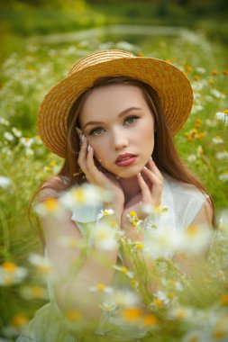 Hasır şapkalı hassas bir kız yeşil çimenler ve papatyalarla çevrili bir şekilde tarlada oturuyor. Yaz güzelliği, makyaj ve çiçekler..